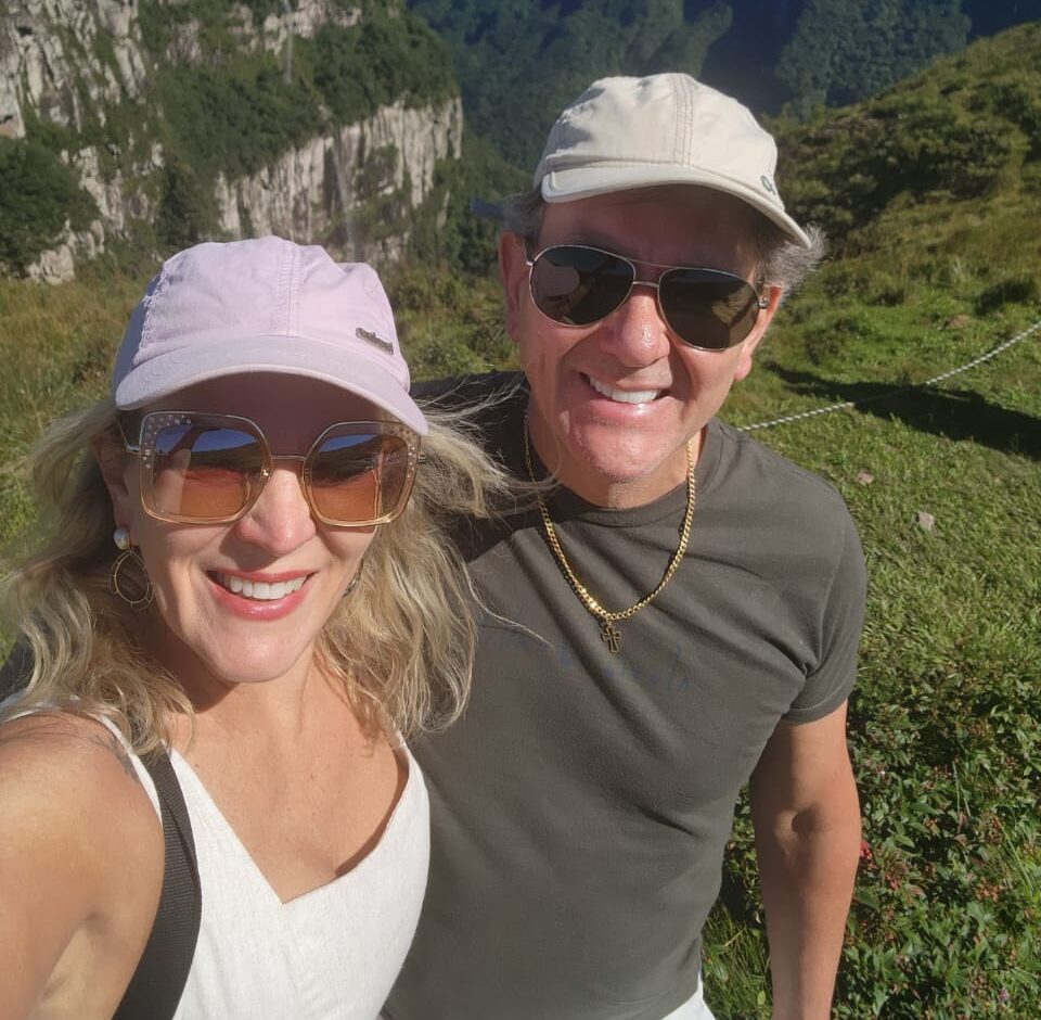 Elaine, autora do Lojas de Esportes, de boné lilás e óculos de sol em uma selfie numa trilha de montanha, ao lado de um companheiro de caminhada